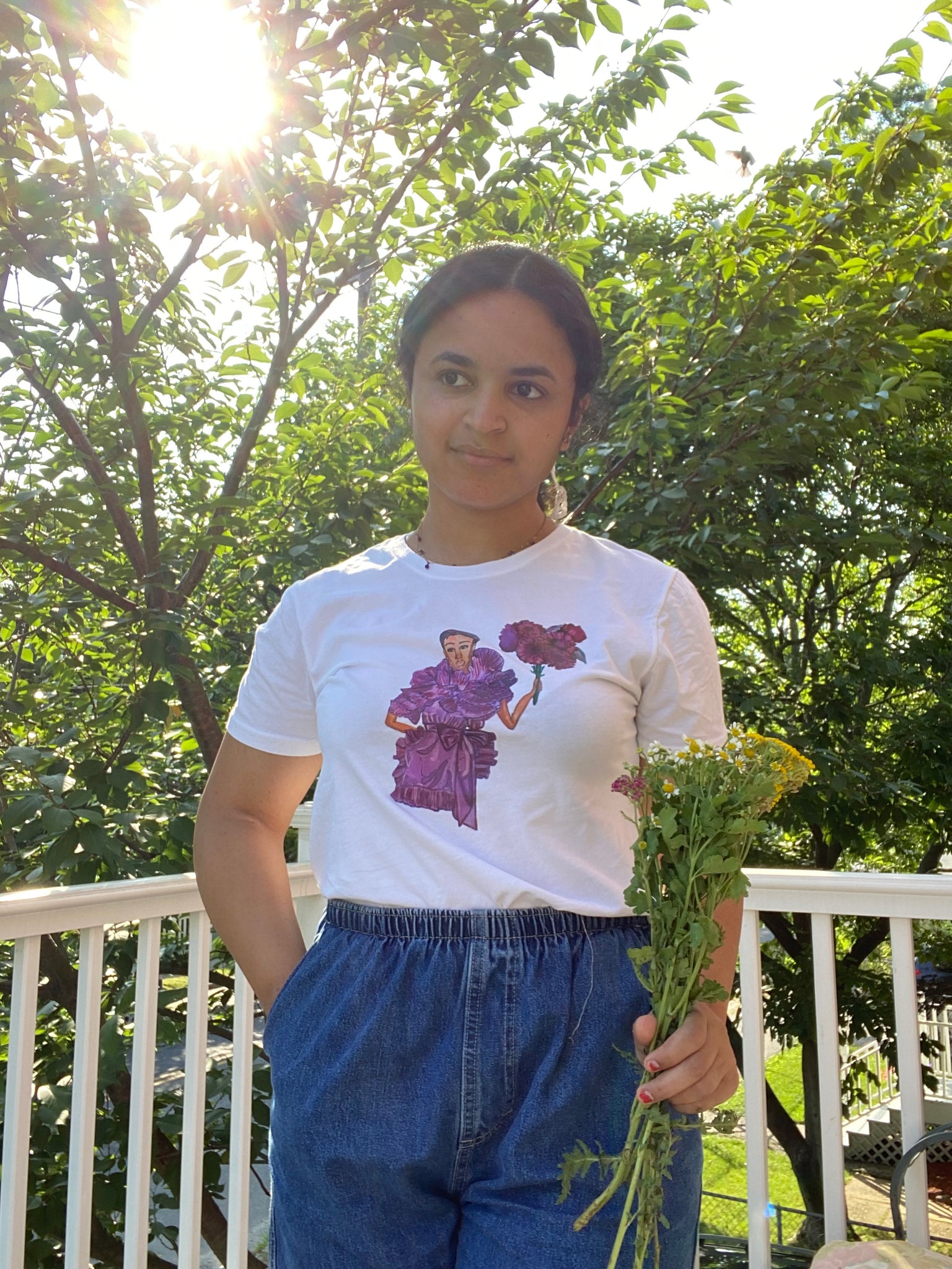 A girl holding flowers and wearing denim shorts and an upcycled white t-shirt with an illustration on it of model Janaye Furman in a purple ruffled gown