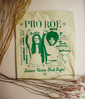 A folded t-shirt with an image on it of Gloria Steinem and Dorothy Pitman and text that reads 'PRO ROE,' surrounded by dry flowers