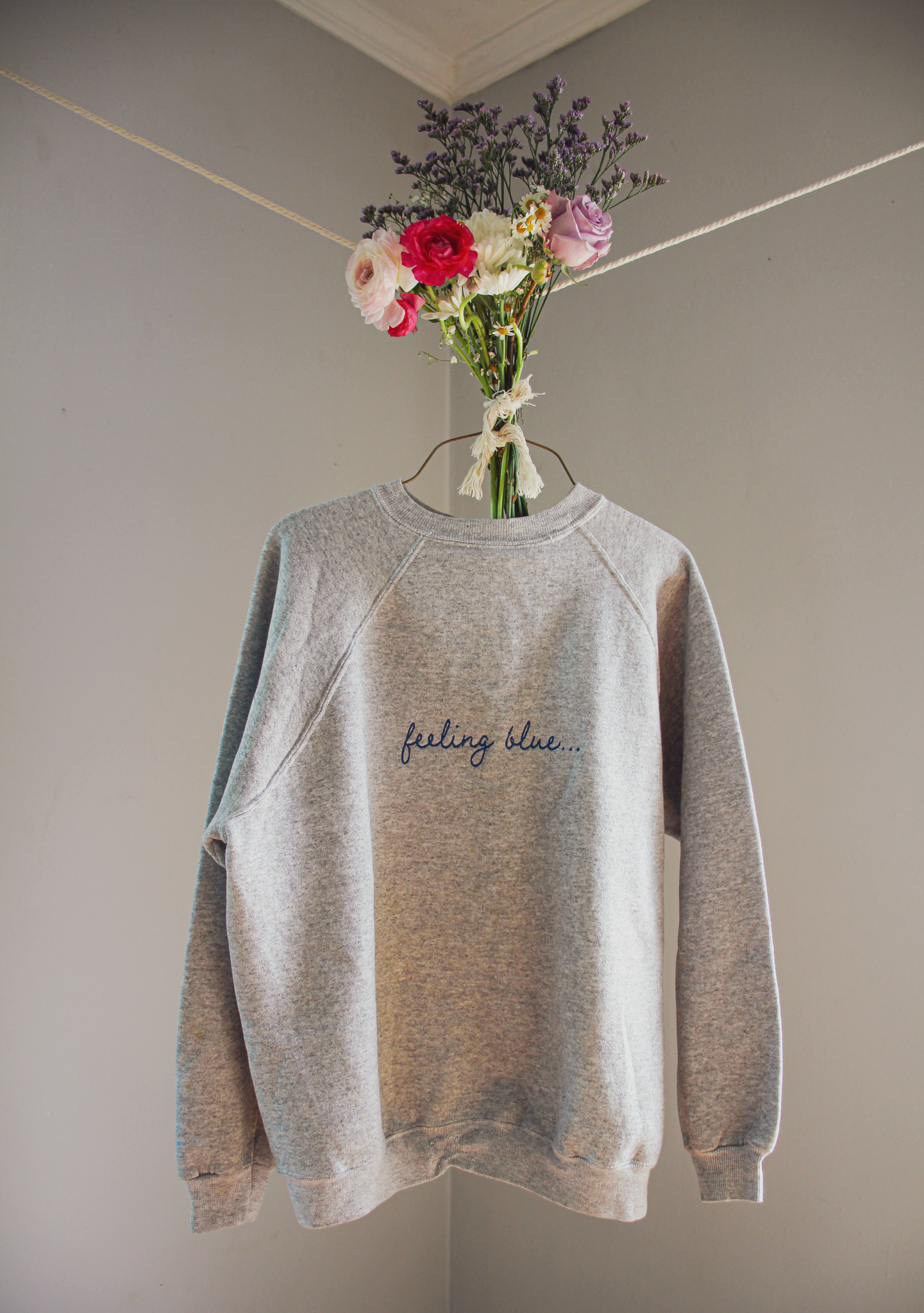 A gray sweatshirt with blue embroidered text on it that reads "feeling blue....," hanging on a wire hanger with a bouquet of flowers tied to it
