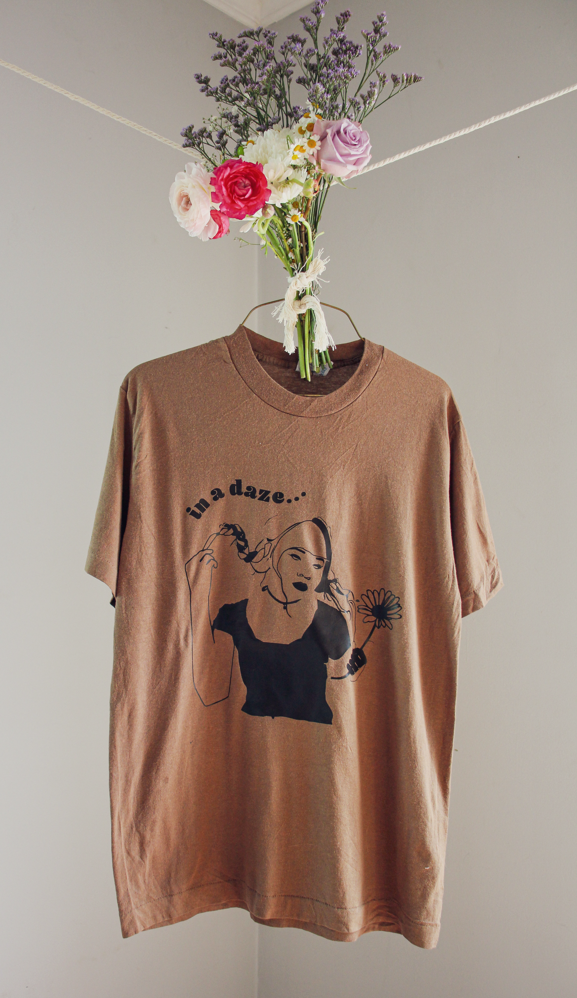 A brown t-shirt with a black illustration of a girl holding a flower and text that reads "in a daze....," hanging on a wire hanger with a bouquet of flowers tied to it