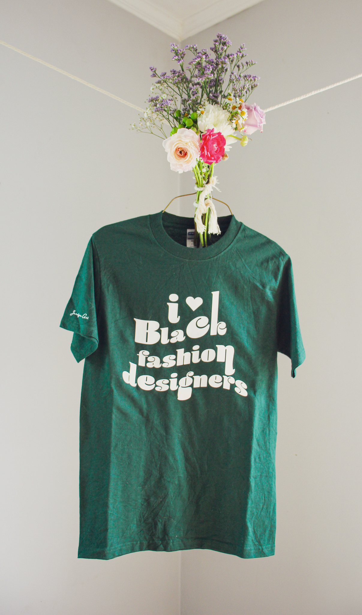 A dark green t-shirt with retro text on it that reads "i love Black fashion designers," hanging on a wire hanger with a bouquet of flowers tied to it