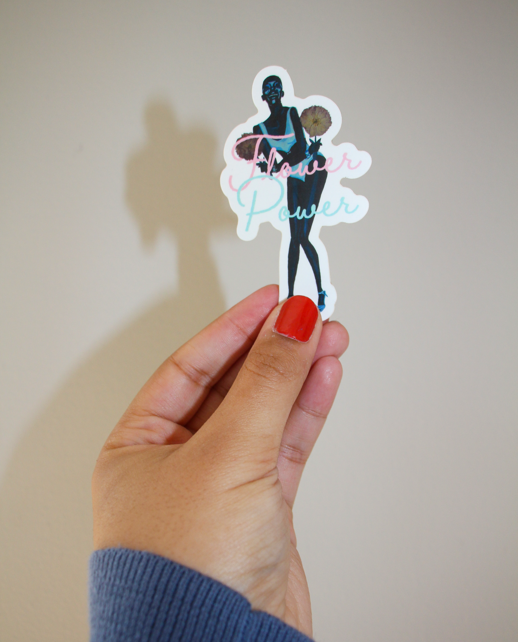 A hand holding a sticker of an image of model Adut Akech holding pressed flowers, with the words "Flower Power" in front