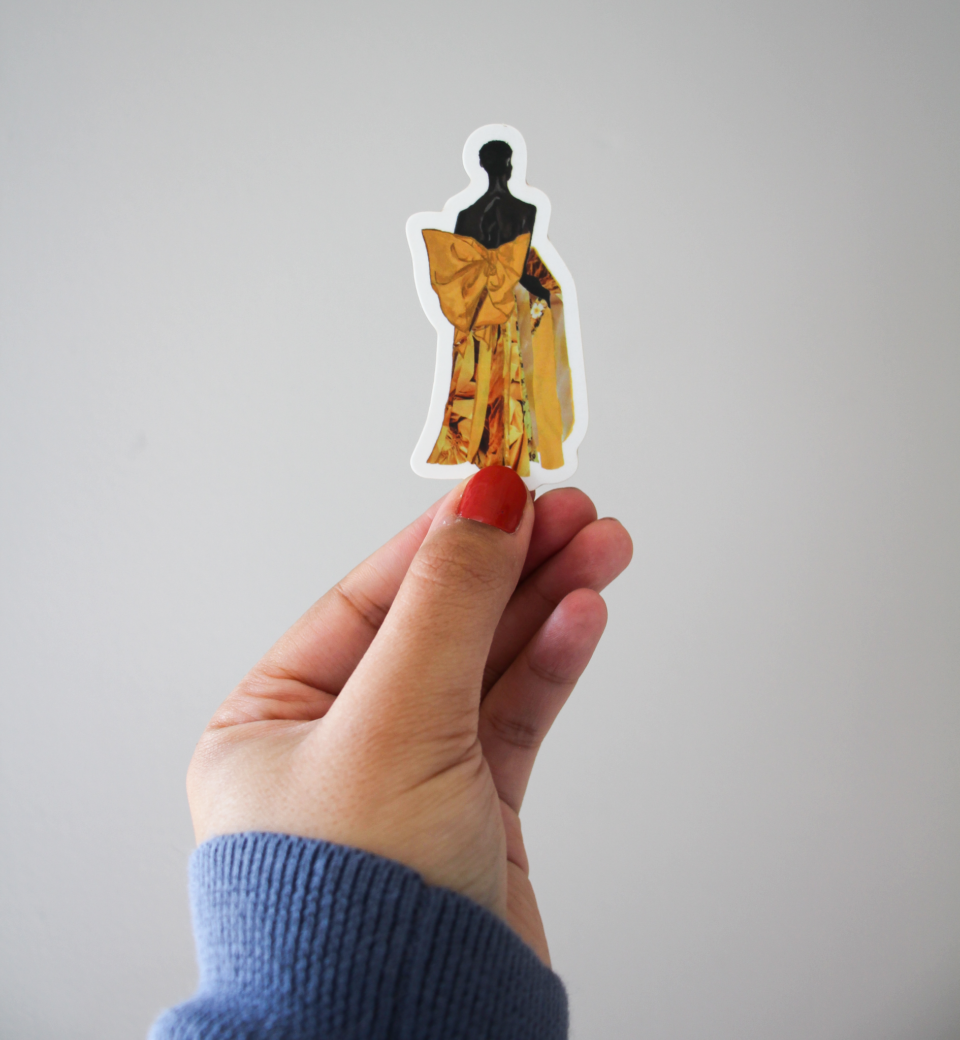 A hand holding up a sticker with an illustration of Black model Akiima Ajak wearing a yellow Valentino gown with a large bow in the back