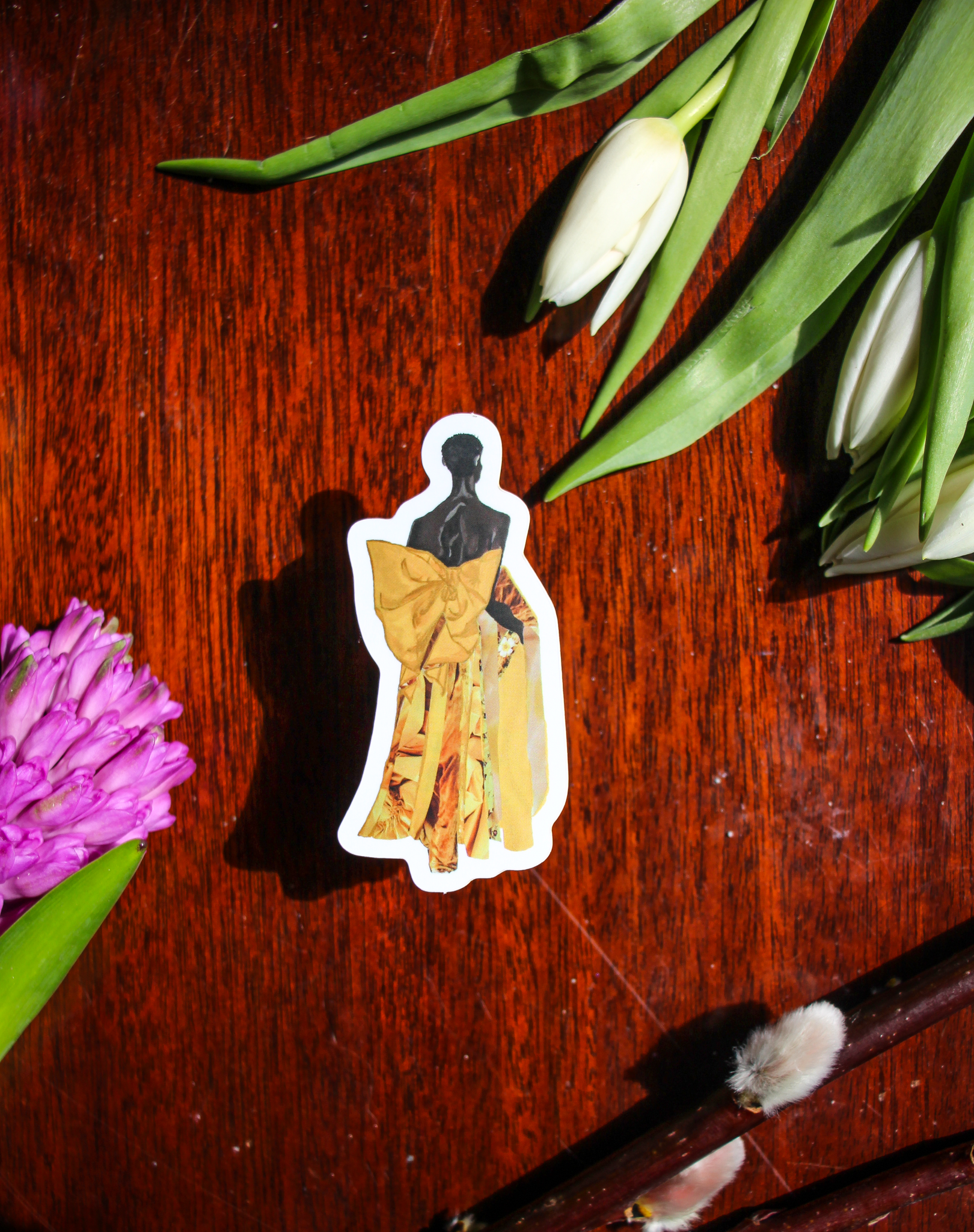 A sticker with an illustration of Black model Akiima Ajak wearing a yellow Valentino gown with a large bow, lying on a wood table surrounded by flowers