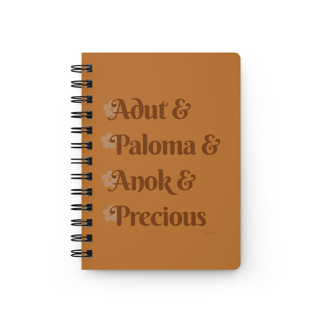 An orange notebook with dark orange text that reads "Adut & Paloma & Anok & Precious" in reference to the Black models Adut Akech, Paloma Elsesser, Anok Yai, and Precious Lee