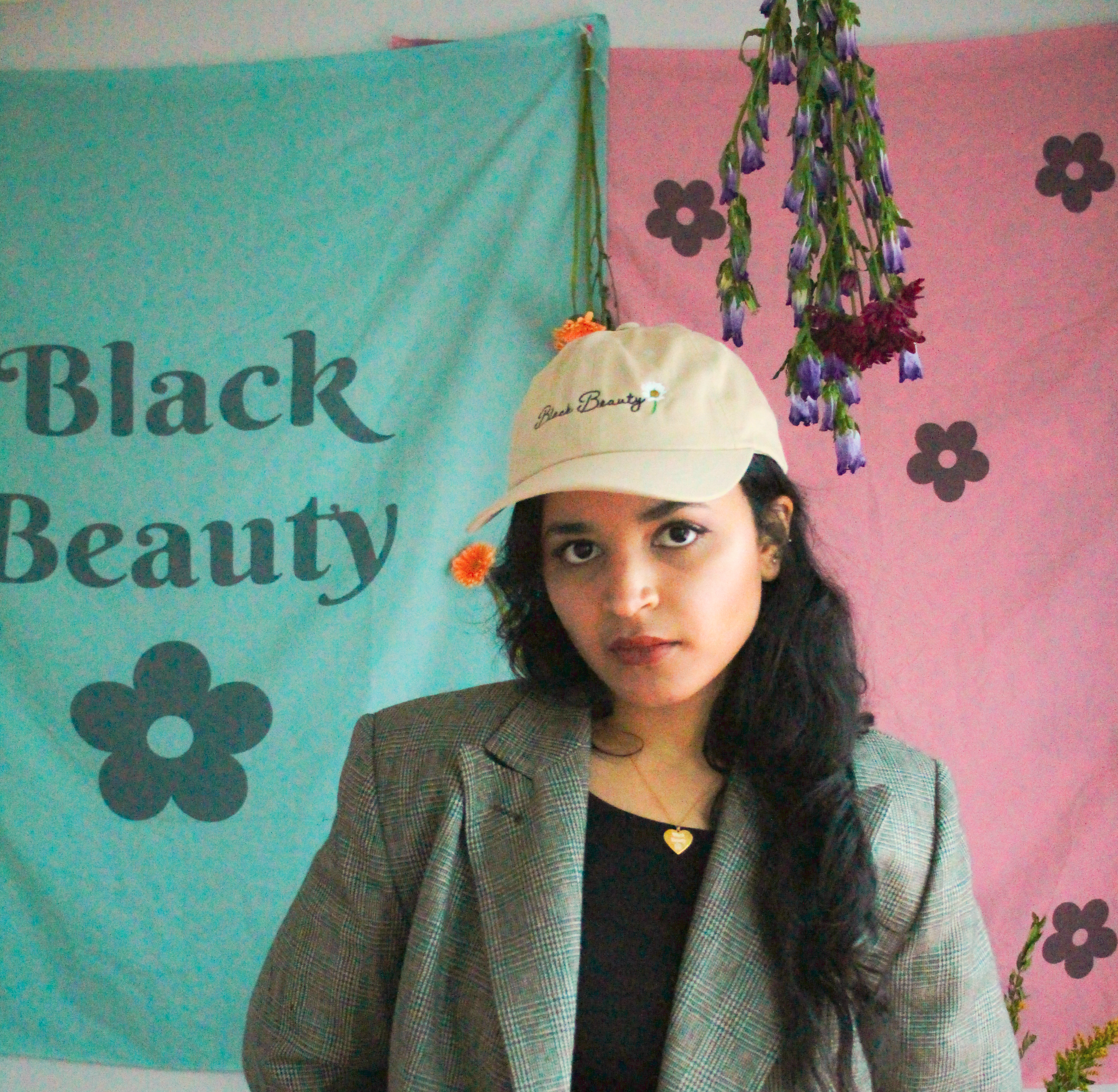 A woman standing in front of green and pink tapestries while wearing a beige baseball cap with embroidered text on it that reads "Black Beauty"