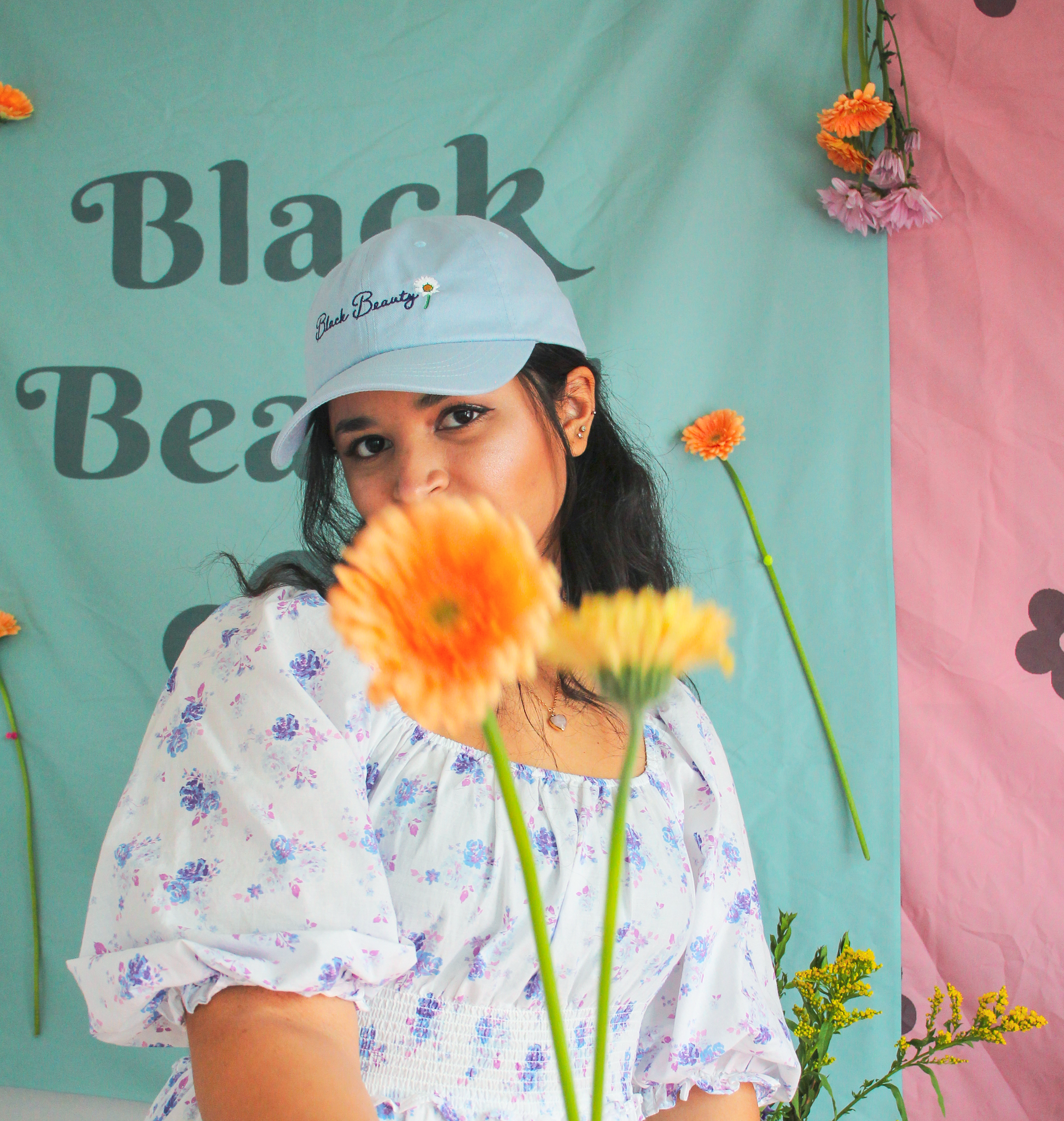 A woman posing with orange flowers and wearing a light blue baseball cap with embroidered text on it that reads "Black Beauty"