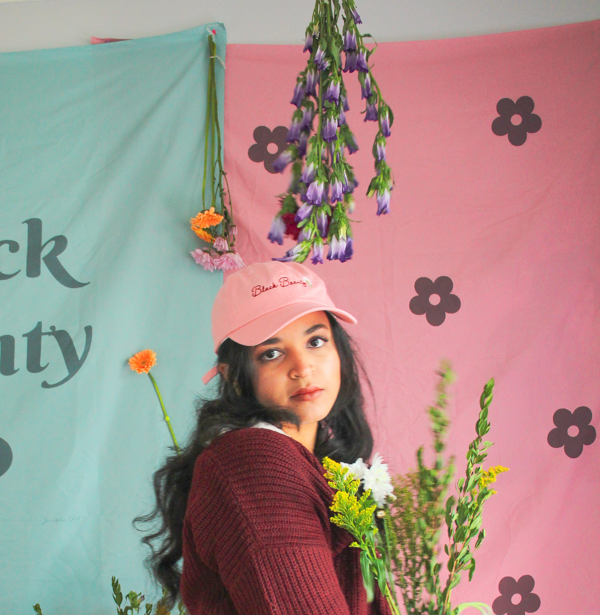 A woman standing in front  of pink and green tapestries while wearing a light pink baseball cap with embroidered text on it that reads "Black Beauty"