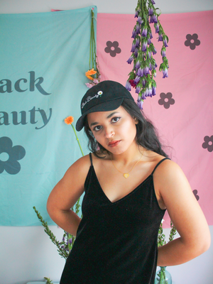 A woman standing in front of green and pink wall tapestries while wearing a black velvet dress and a black baseball cap with embroidered text on it that reads "Black Beauty"