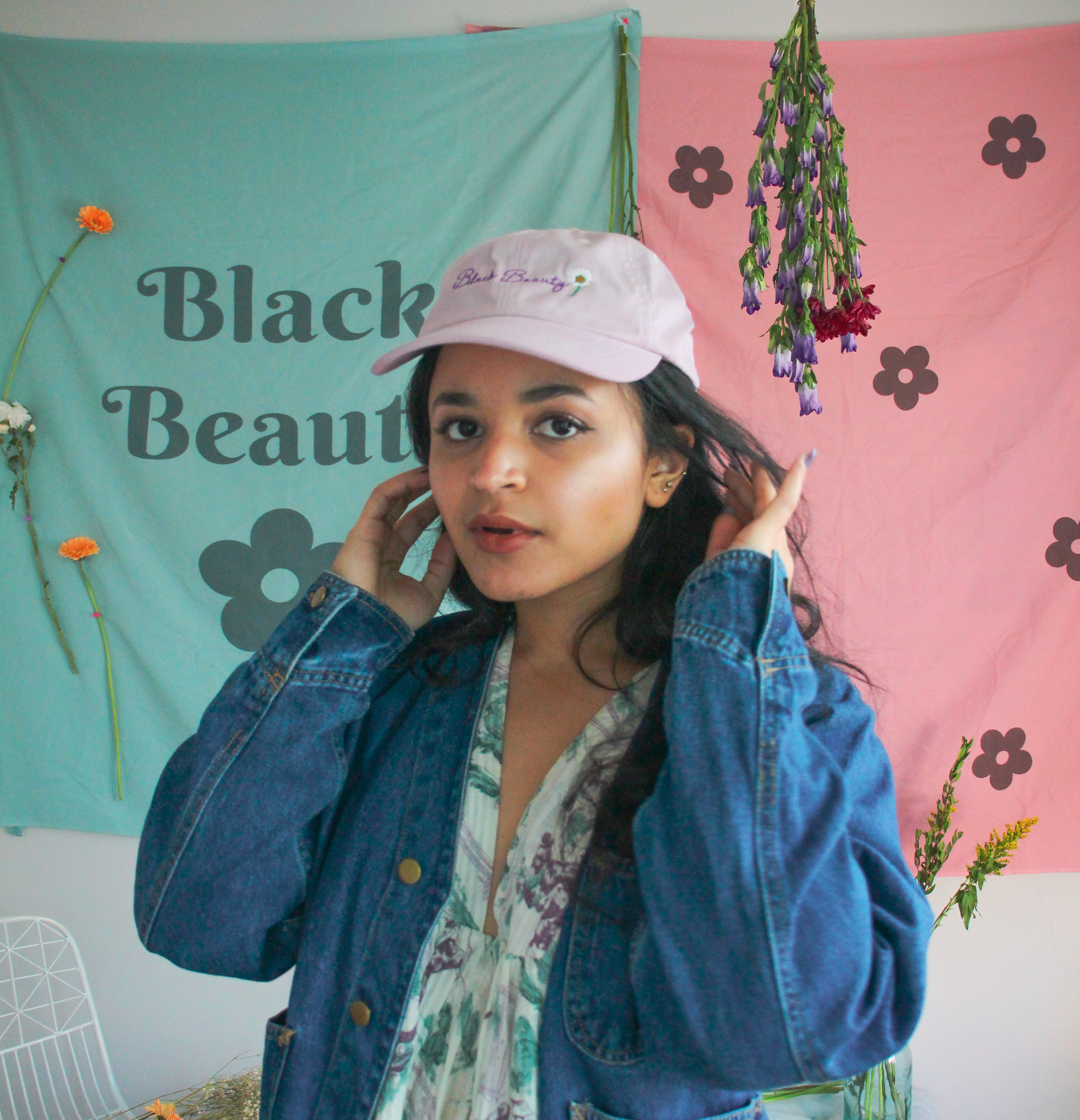A woman posing in front of green and pink wall tapestries while wearing a lavender baseball cap with embroidered text on it that reads "Black Beauty"