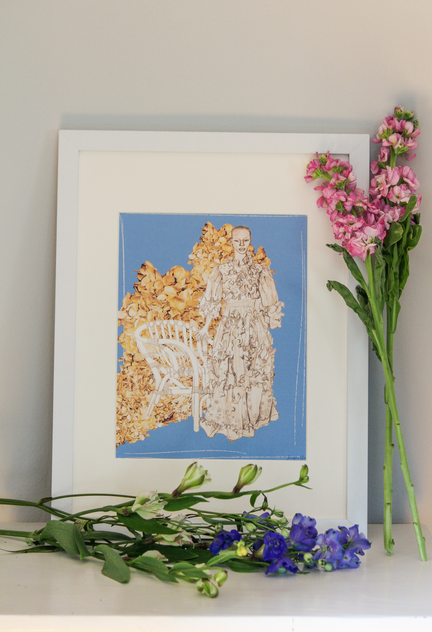 An illustration print with a light blue background and a drawing of Black model Shanelle Nyasiase posing next to a whicker chair in a long Valentino ruffled prairie dress, collaged with cut-out images of dried brown flowers, inside of a white frame that is standing up on a table next to pink and blue flowers