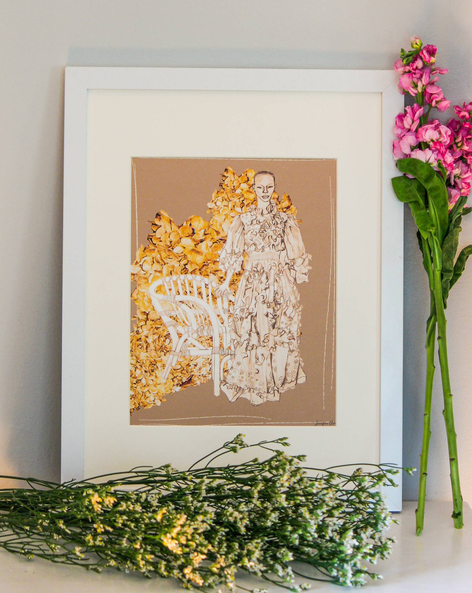 An illustration print with a light brown background and a drawing of Black model Shanelle Nyasiase posing next to a whicker chair in a long Valentino ruffled prairie dress, collaged with cut-out images of dried brown flowers, inside of a white frame that is standing up on a table next to pink and white flowers