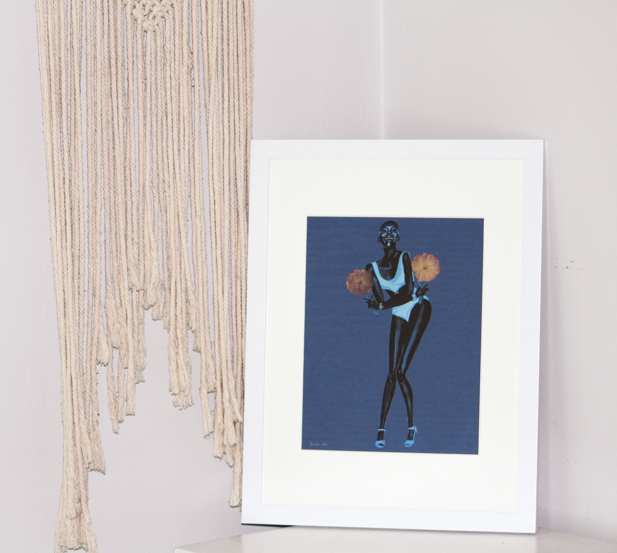8.5 by 11 inch Black fashion art print of a painting of Black model Adut Akech wearing a Chanel one-piece swimsuit and holding pink pressed flowers with a dark blue background in a white frame with matting, standing up on a white table with tassel wall decor behind it 