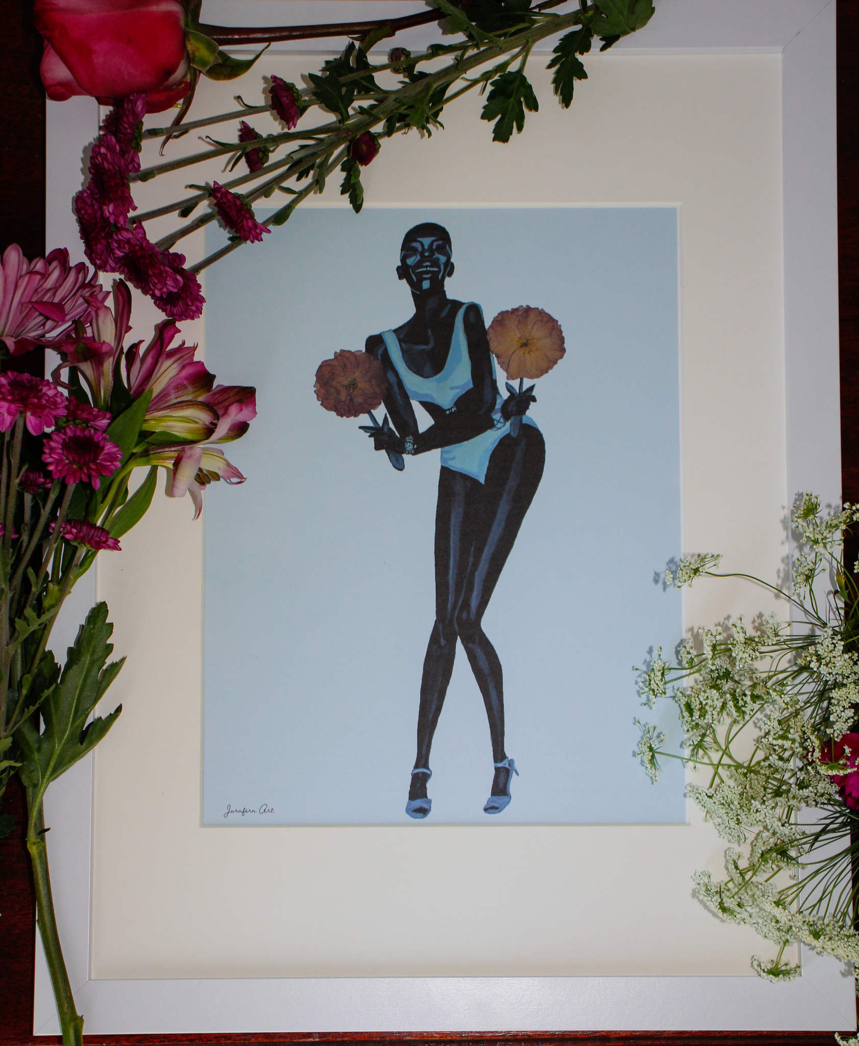 8.5 by 11 inch Black fashion art print of a painting of Black supermodel Adut Akech wearing a Chanel swimsuit and holding pink pressed flowers with a light blue background, in a white frame with matting lying on a table surrounded by fresh pink flowers