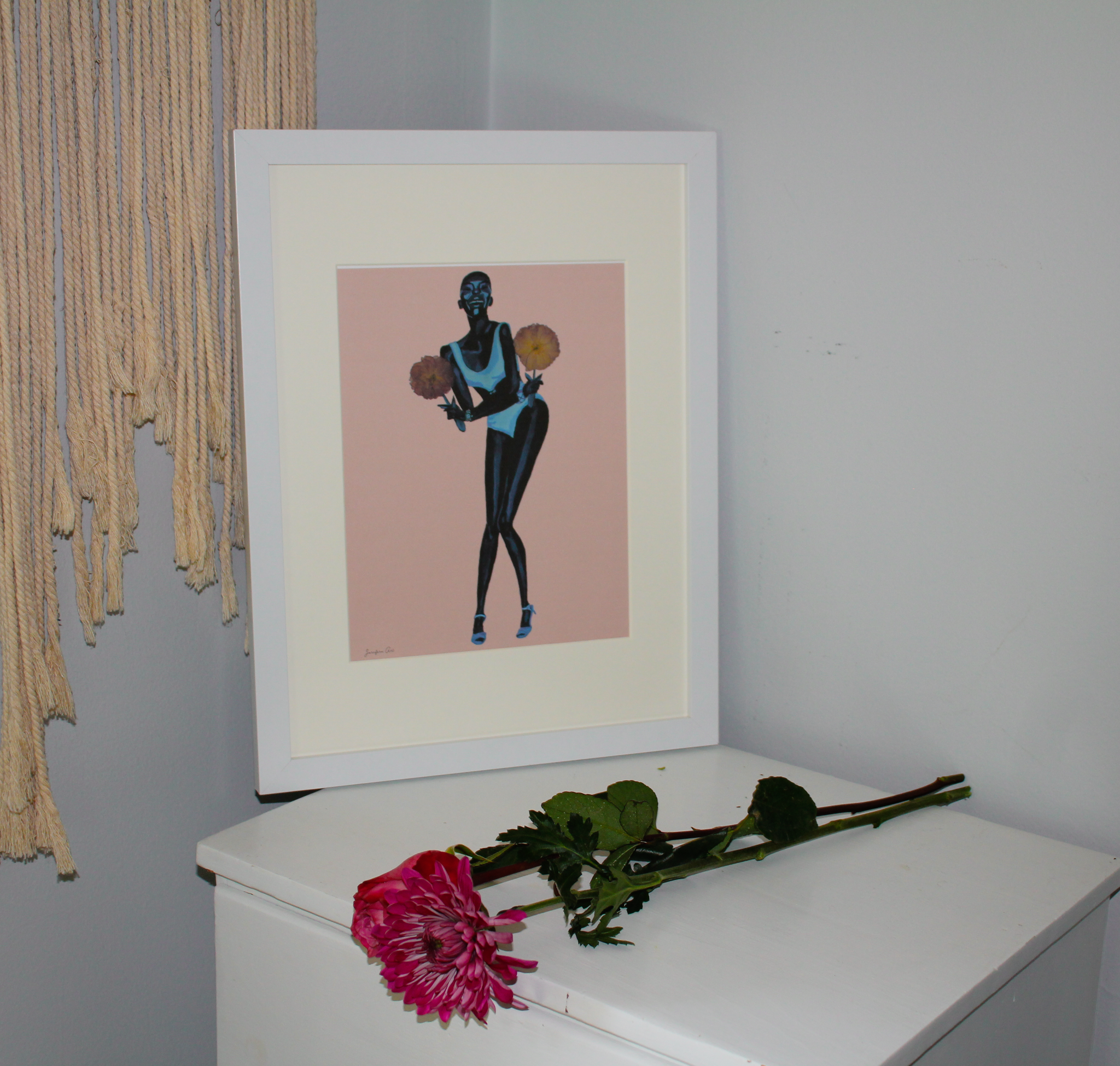 8.5 by 11 inch Black fashion art print of a monochrome blue painting of Black supermodel Adut Akech wearing a Chanel one-piece swimsuit and holding pink pressed flowers with a light pink background in a white frame with matting, standing up on a white table with pink flowers lying next to it