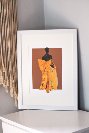 A framed illustration print with a brown background and model Akiima Ajak wearing a yellow Valentino gown with a large bow in the back, resting on a white table with tassels hanging behind it