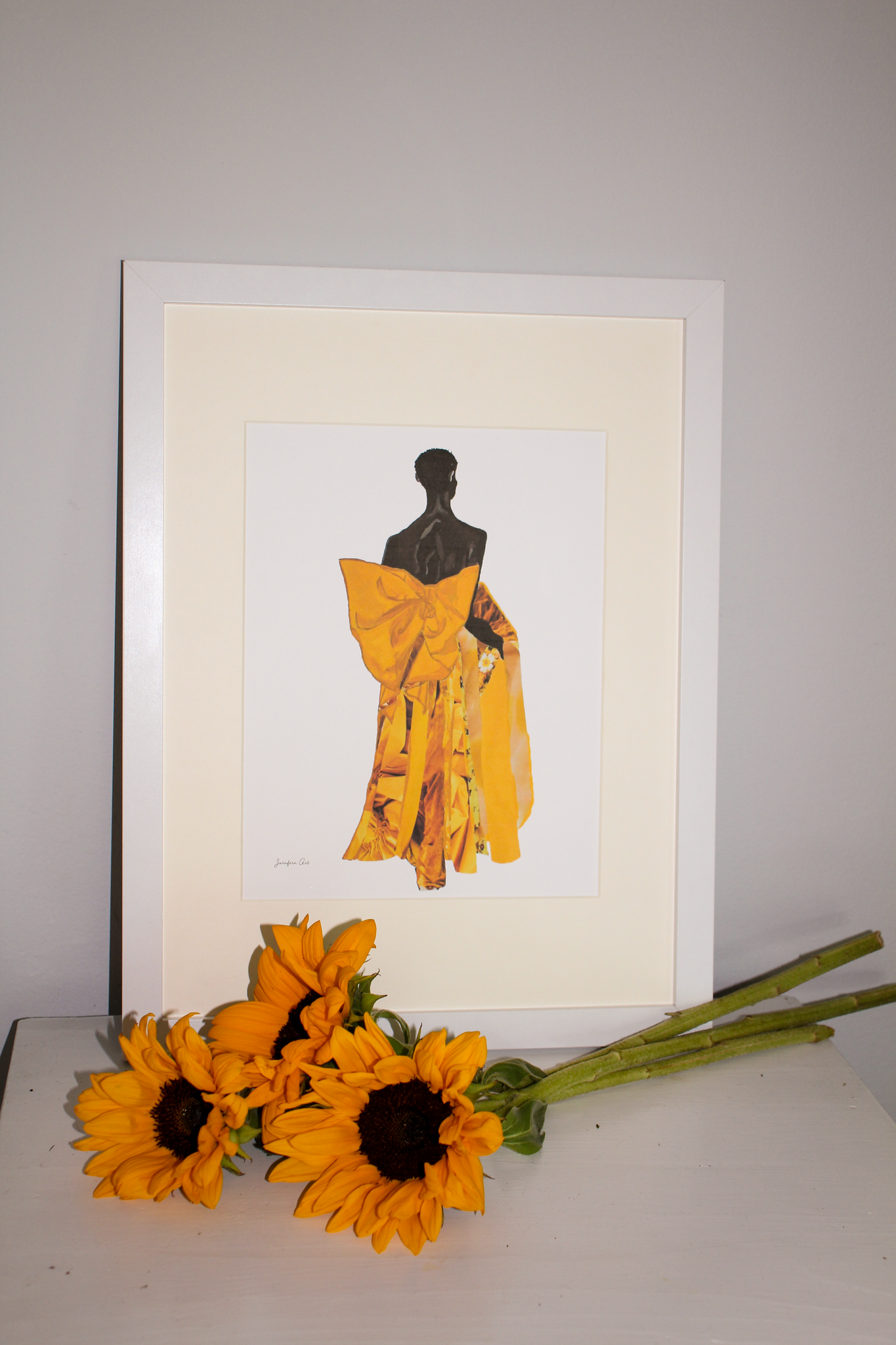 A framed illustration print of model Akiima Ajak wearing a yellow Valentino gown with a large bow in the back, and a white background, displayed on a table with sunflowers
