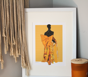 A framed illustration print with a yellow background and model Akiima Ajak wearing a yellow Valentino gown with a large bow in the back, displayed on a table next to an orange candle and tassel wall decor 