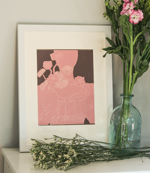 A monochrome pink graphic illustration of a woman in a top with bows holding a vase of flowers, with a brown background, inside of a white frame standing up on a table next to a vase of garden flowers and bunch of loose white flowers