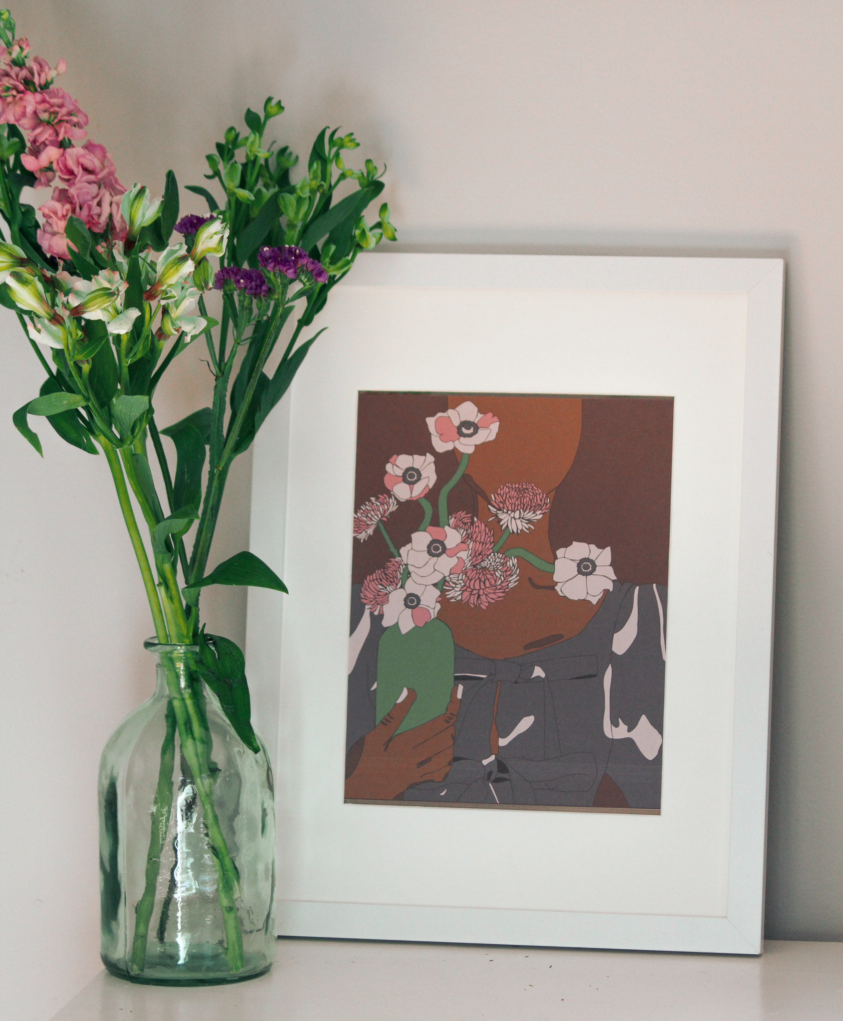 A graphic illustration of a Black woman in a black top holding a green vase of pink flowers, with a brown background, inside of a white frame standing up on a table next to a vase of garden flowers